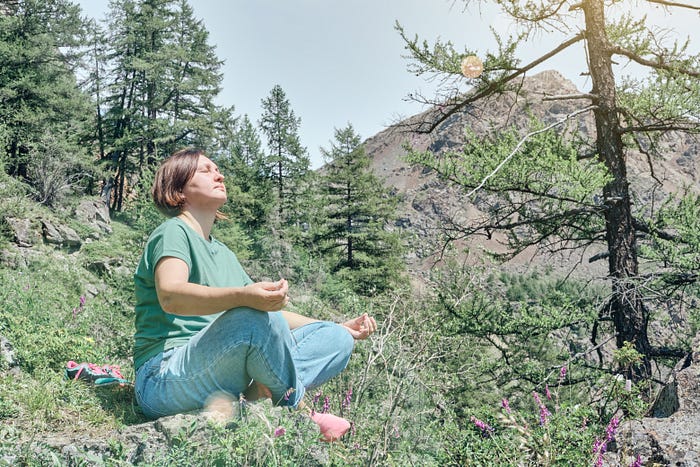 A woman sitting cross legged in the woods, near the top of a hill meditating in the sunlight, this image shows size inclusivity of people of all sizes, content relevant to body mass index (BMI), level of fitness, health, wellness, exercise, nutrition, healthy living, healthy lifestyle, weight stigma, anti-fat phobia, new norm, healthy weight