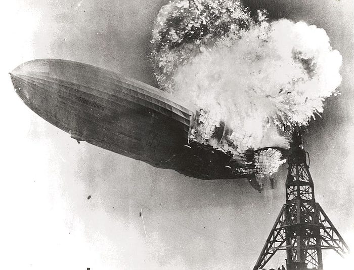 How Are Big Airships Really Making A Comeback Again? — An image of an awesome burning Hindenburg near some sort of an electricity tower. The rear end of the airship is seen to be releasing a massive ball of flame that is 4–5 times the diameter of the airship itself.