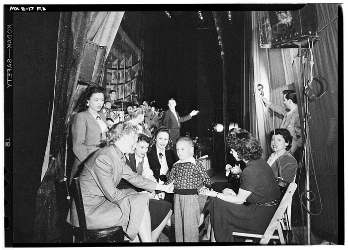 A black-and-white photograph of American singer June Christy being amongst other women backstage, interacting with a child. Behind them features a music ensemble.