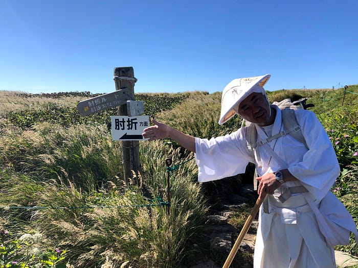 Master Kazuhiro Hayasaka of Daishinbo pilgrim lodge in front of a sign pointing out the routes up Mt. Gassan of the Dewa Sanzan