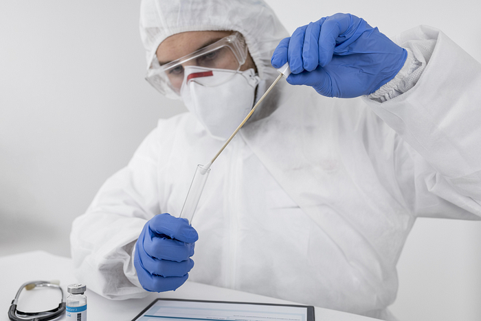 A person wearing a mask, gloves, and protective clothing inserting a nasal swab into test tube.