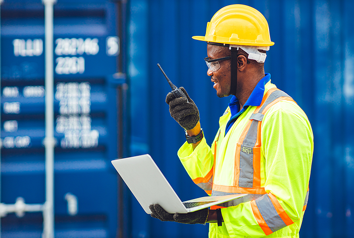 Worker at a cargo port using 2-way radio and holding a laptop.