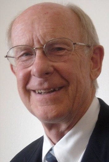 Dr. Johnston sits in front of a white background and is wearing a dark suit and grey-rimmed glasses.