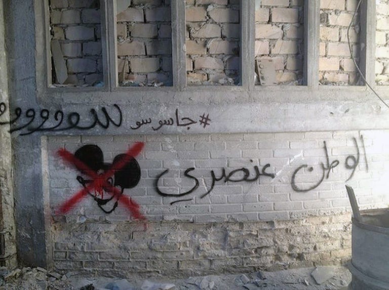 Photograph showing an Arabic script on a wall in Syria alongside a graffiti of a Mickey Mouse character’s head with a red cross on top of it.