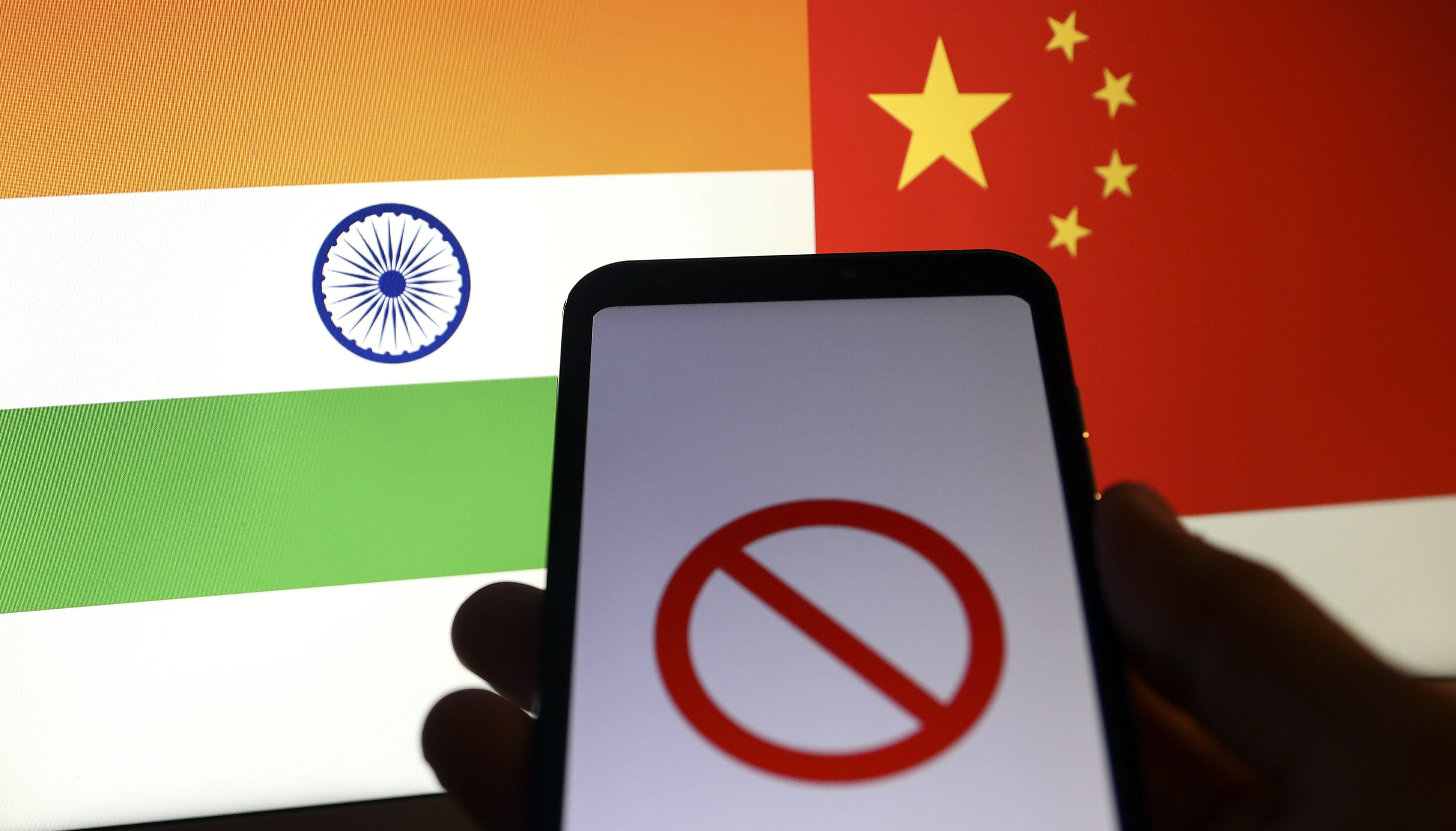 Indian flag and Chinese flag are displayed on a laptop screen behind a smart phone displaying a symbol of “block”