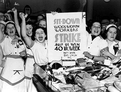 Women store clerks raise their fists and raise a sit-down strike sign demanding a 40-hour work week for Woolworth workers.