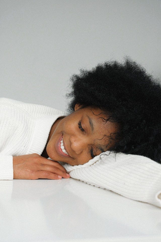 A woman smiles, eyes closed and rests her head on a table.