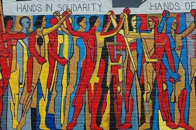 A brick wall painting of hands in solidarity. Abstract nude persons clasping hands with red, blue, yellow, black, and brown.