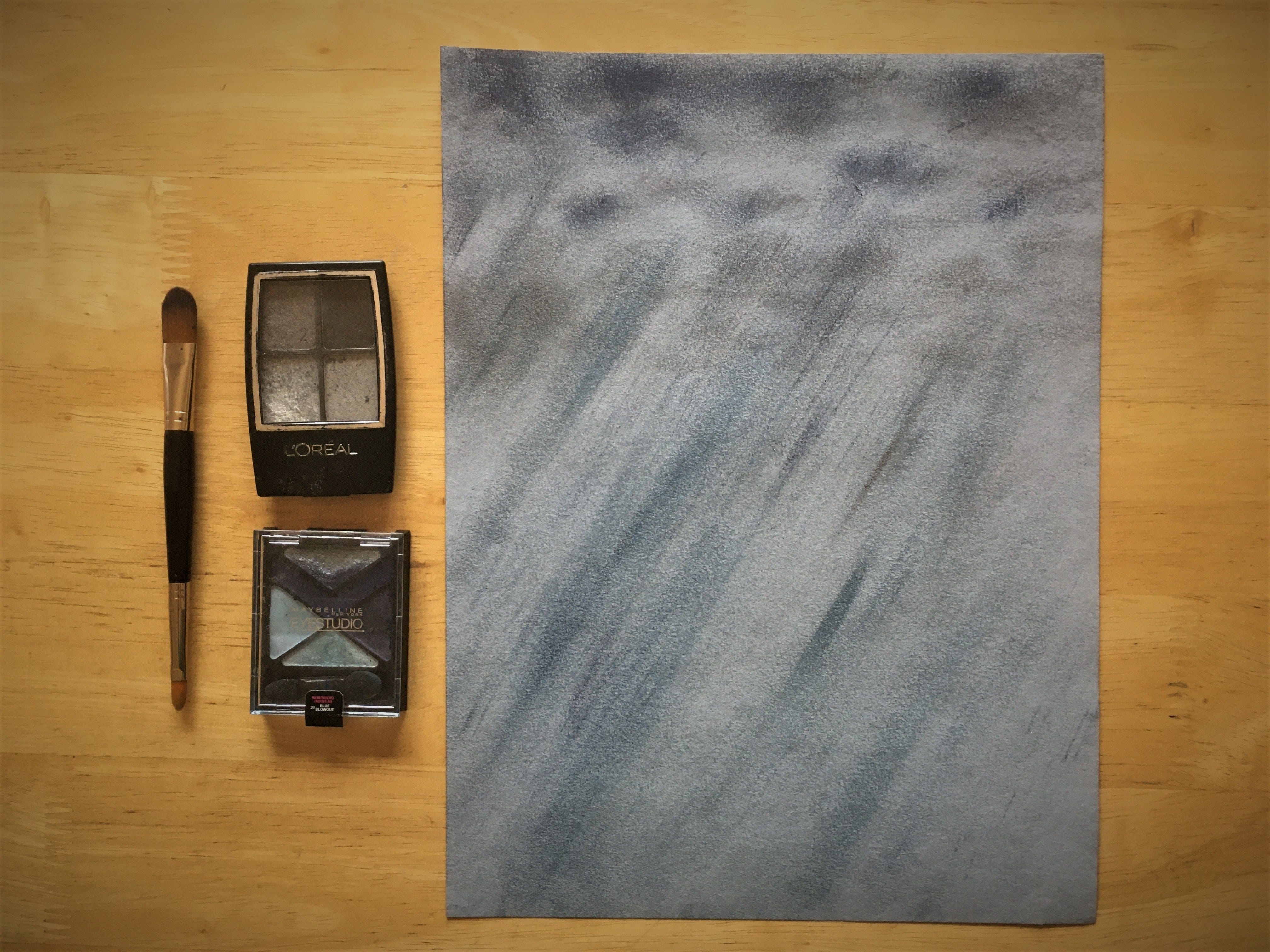 Eyeshadow palettes sit alongside a “drawing” of gray clouds and driving blue rain, created with the makeup.