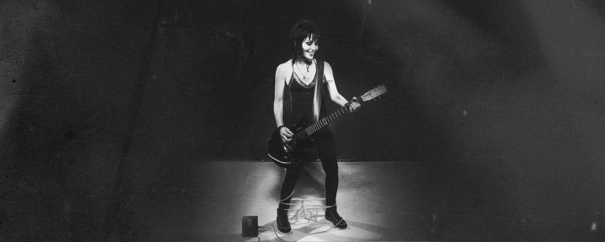 Joan Jett on stage with a guitar and cords wrapped around her ankles. Black and white photo.