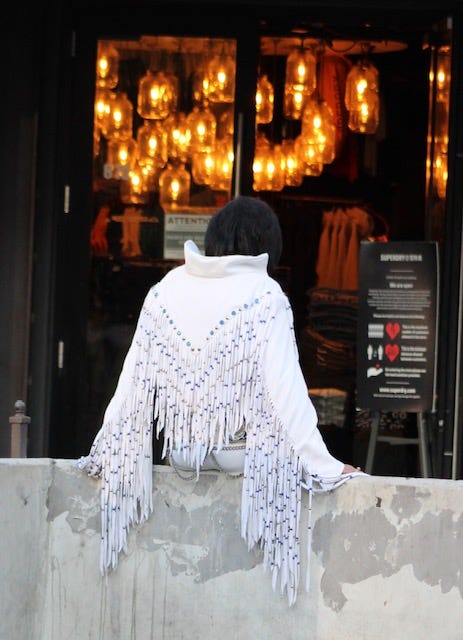 photo of an Elvis impersonator sitting on a roadside block on the Las Vegas strip in a fancy white jacket with long tassels and high collar, with his back to the camera