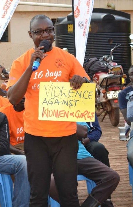Emmanuel Mensah wearing an orange t-shirt, holding mic on the right hand and a yellow coloured sign that has a writing of: ‘VIOLENCE AGAINST WOMEN AND GIRLS’ on the left hand