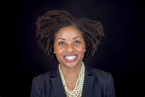 Dr. DeJarnett has short dark hair and sits in front of a black background, wearing pearls and a dark blue suit.