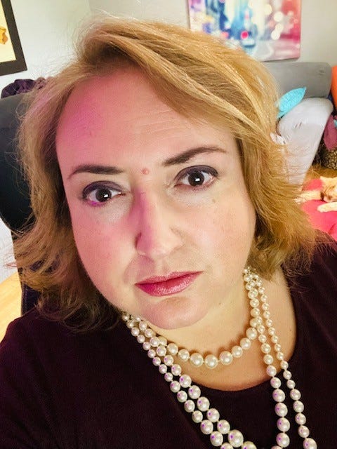 Melissa Parker, a woman who has shoulder length sandy-blond hair, brown eyes, and pearl necklaces. She looks at the camera with a neutral expression. In the right-hand corner of the photo, we can see the head and two paws of her orange cat.