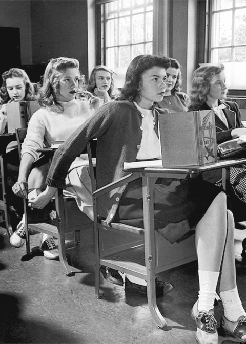 A black and white photo of a classroom in 1944 where two girls are passing notes