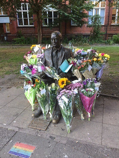 A statue of Alan Turing sitting on a bench surrounded by bouquets of flowers