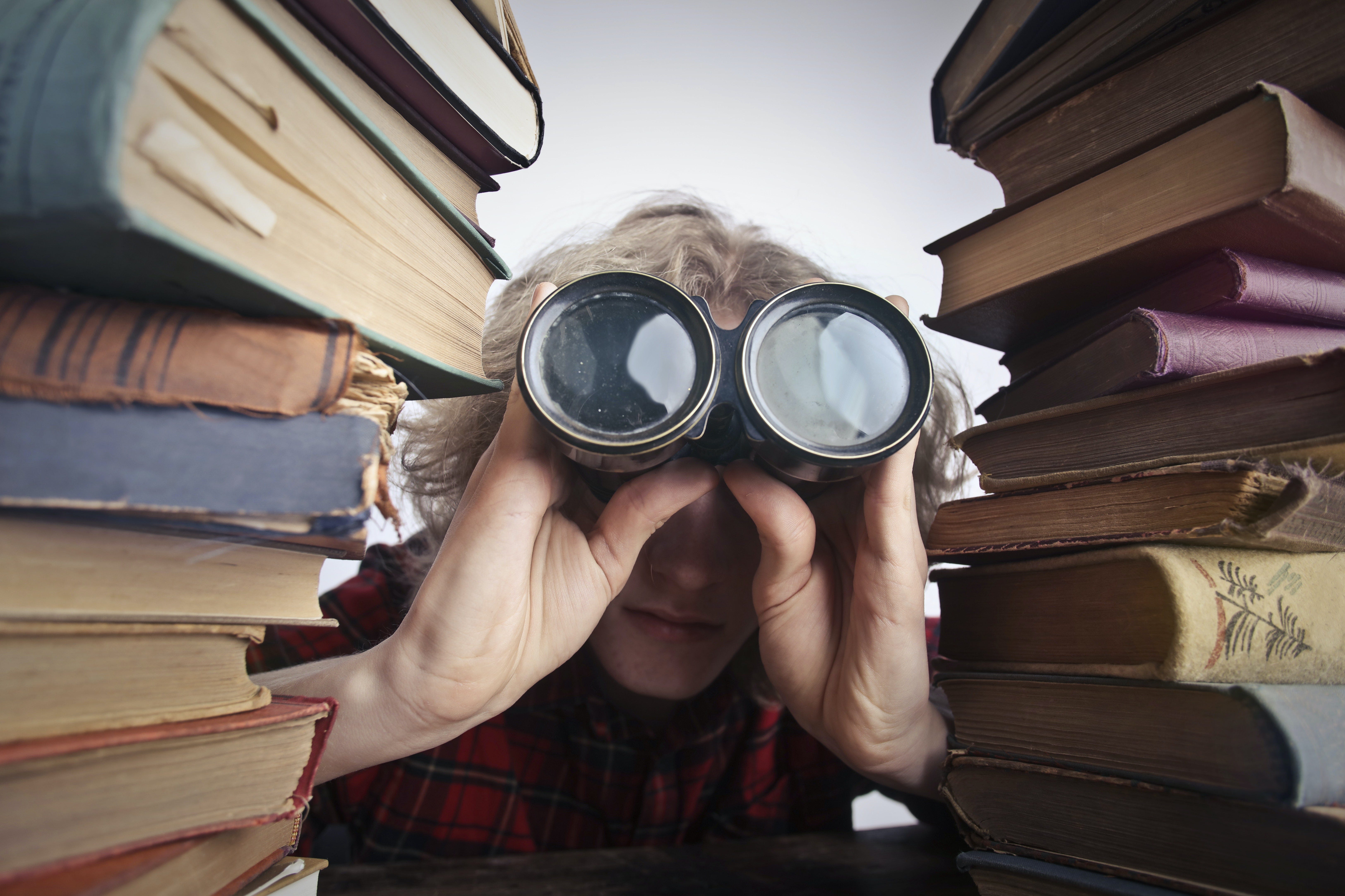 A woman holds binocular lenses between two piles of books.