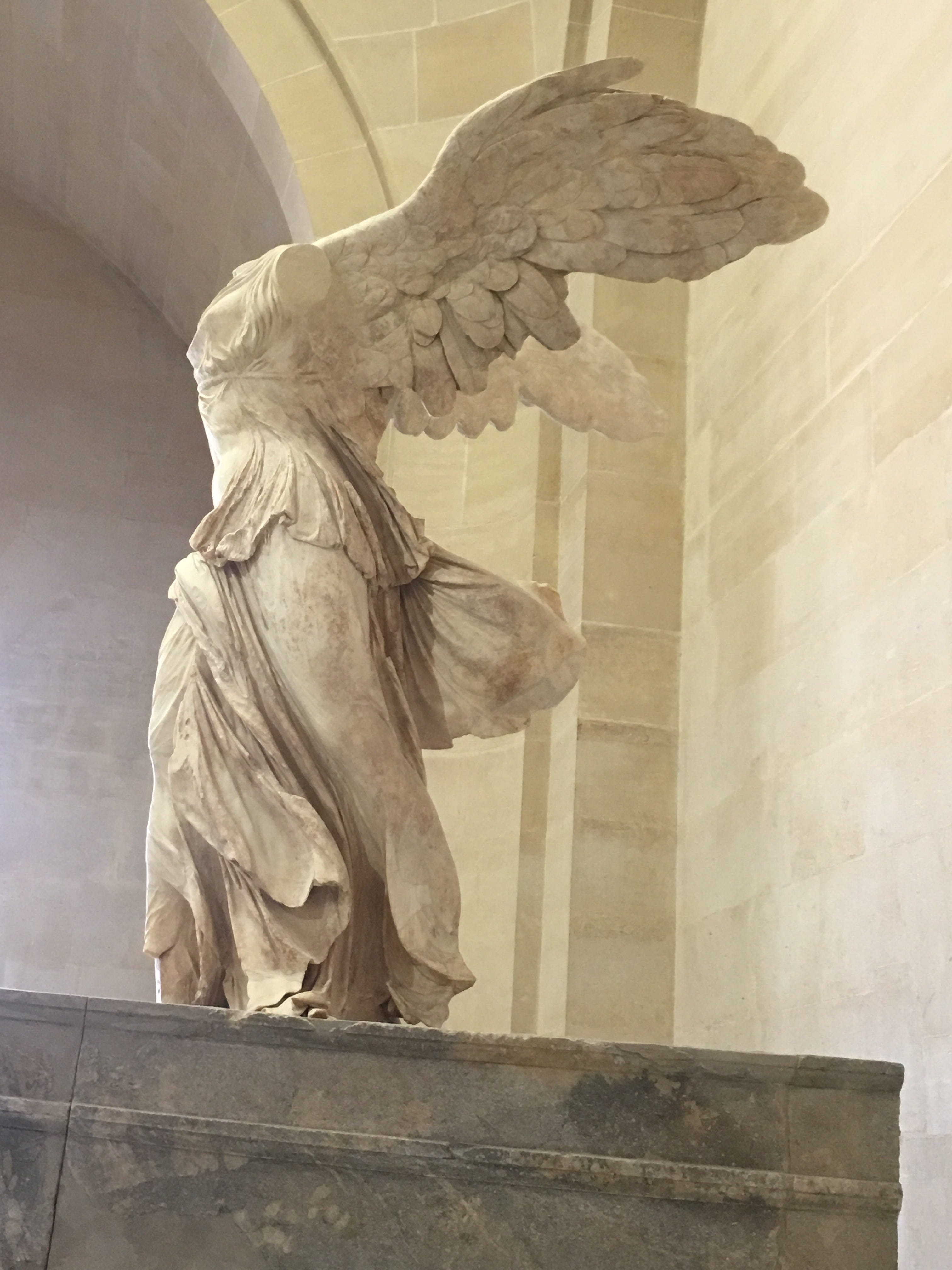 A woman with outstretched wings is standing on the prow of a ship. Her garments are twisting and billowing in the wind.