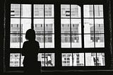 A silhouette of a person standing by a large window, looking out at a building with multiple windows on the opposite side. The scene is captured in black and white, highlighting the contrast between the dark interior and the bright exterior. The person appears contemplative, adding a sense of introspection and solitude to the image.