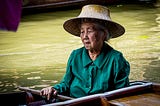 An elderly East Asian woman sits in a boat on a green river. She stares thoughtfully downward, as if she is remembering something.