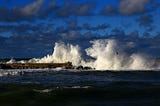 An explosion of breaking waves overwhelms a break water. A lone seagull flies low before the white spray. The sky is blue with scattered clouds.