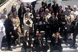 A photo of a large group of Good Omens cosplayers. They are mostly Crowley and Aziraphale cosplayers with a few other characters as well. No two of them look the same, despite the fact that most of them are cosplaying the same two characters.
