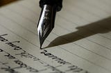 This is a spotlight of a calum calligraphy silver pen head writing on thin rice paper, which is an art form. Its shadow is angled to the right, its head upright with ink flowing perfectly from the pen tip, and it is writing out beautiful penmanship. The view of the words is upside down in the image and angled in the lower left corner.