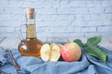 clear glass bottle with apple cider vinegar beside sliced apples on blue cloth