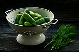 A metal colander on a black surface with a black background. Inside, a pile of small, bright green cucumbers, next to it, a dark green bunch of fennel.