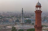 A picture showing two iconic landmarks in Lahore the “Minar-i-Pakistan” (Tower of Pakistan) and a minaret of the Badshahi Mosque with many small buildings scattered behind them. The sky is somewhat smoky and dusty from pollution.