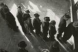 Black-and-white photo showing several people standing in a line for work, viewed from above. They wear hats and coats and appear to be waiting near a doorway.