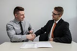 Two men in suits shake hands in an interview-style room.