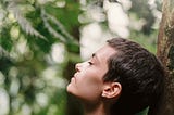 Person leaning their head back against a tree with eyes closed. They are feeling the sun on their face and look relaxed and at ease in the forest around them.