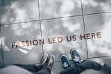 Two pairs of legs and shoes are shown from above as two people stand side by side looking down at large paving tiles and words that read “passion led us here”