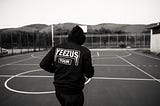 A black and white picture of a man walking or jogging away on a basketball court. He is wearing a hoodie that says YEEZUS Tour on the back.