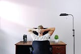 A guy with his hands crossed behind his head, sitting in front of a desk with a monitor.