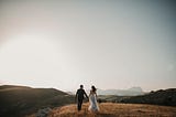 A photograph of a bride and groom on a mountain, holding hands and looking into the distance