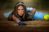 Girl with a crown on her head touching a softball base with a yellow softball nearby.