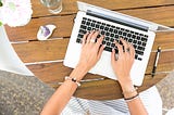 image shows a woman’s hands hovering over a silver laptop on a wooden table