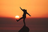 A figure in silhouette stands on a rock; the sun sets behind them; they stand balanced on one leg, the other leg, their arms stretched out.