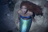 A young Black woman with red locs, actress Halle Bailey, sits on a rock underwater. She is wearing a purple bustier and has a shimmering green mermaid tail. She is looking up, and her mouth is open in song.