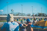 Race fans at a speedway cheering on race cars as they round the track coming out of a turn into the straightaway