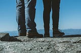 A couple stands on a mountain peak looking across the horizon. Only their legs and feet are shown atop the granite