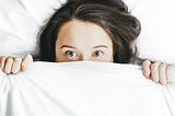 A terrified woman, with a white bedsheet pulled over her eyes. You can see her brunette hair, her face upto her eyes, and two hands clutching the bedsheet.