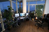 Picture of an office, desk in front of window, three computer monitors on.