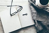 A black and white image of an open book with glasses and a key on it, and next to it a cloth and a cup of coffee.