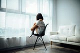 AI Generated photo of a girl sitting on a chair and looking out a window, she has short hair and the room is an unsurprising color of white. She is also wearing white. Kind of boring to look at, really