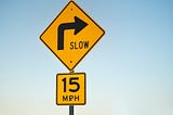 Two road signs stacked on one another indicate a speed limit of 15 miles per hour around a corner. The only copy that accompanies the arrow icon is “Slow” and “15 MPH.”
