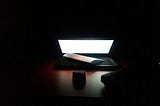 A laptop is half folded, the light of its screen lights up the keyboard. The room is dark and there is a folded letter on top of the laptop’s keyboard. In the dim light emanating from the computer we can see a small speaker.