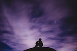 A twinkling night sky with a purple hue, as a silhouette of a small figure sits down; looking up, pondering.